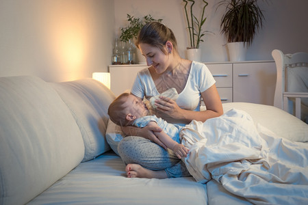 年轻母亲晚上用瓶子在床喂婴儿的肖像图片