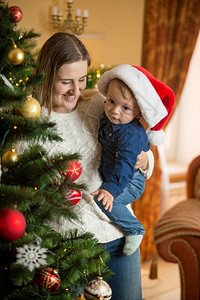 快乐的年轻母亲肖像在客厅圣诞树的上抱着她的婴儿男孩在圣诞树的上在客厅里图片