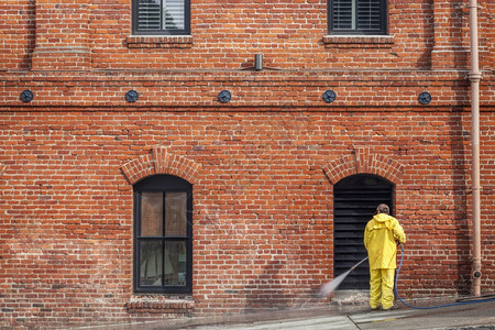 一名城市工人穿着黄雨大衣在砖房前面用水管打人行道图片