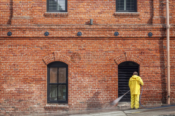 一名城市工人穿着黄雨大衣在砖房前面用水管打人行道图片
