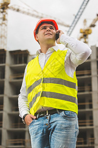 年轻笑的工程师身穿硬帽和安全背心在建筑工地通过电话交谈图片