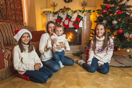 快乐的年轻母亲和孩子们坐在壁炉的地板上在背景装饰圣诞树图片