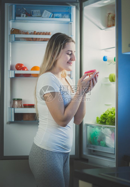 美丽的女人晚上从冰箱吃甜圈图片