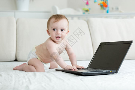 尿布中的婴儿男孩爬在床上并使用膝型电脑图片