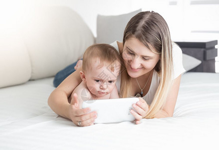 美丽的年轻母亲与婴儿躺在床上并使用平板电脑图片