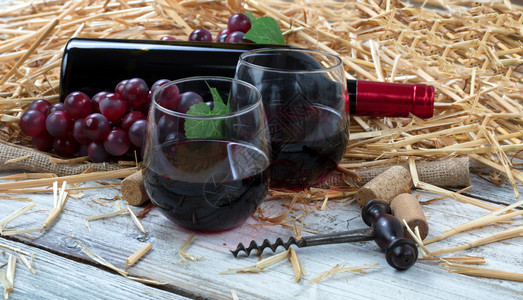 近距离观看装满红酒瓶和葡萄的杯子酒加上装有稻草和白生锈板上布的软木塞图片