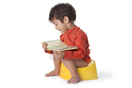 托德勒男孩坐在一个便壶上阅读一本关于白背景的书图片