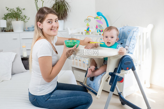 微笑着的年轻母亲用高椅子喂养她幼儿的罪孽并秘密地看微笑着的年轻母亲用高椅子喂养幼儿的罪孽并秘密地看着她们图片