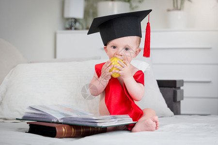 穿着毕业帽子的可爱男孩坐在床上有书和咬苹果穿着毕业帽子的可爱婴儿坐在床上有书和咬苹果图片