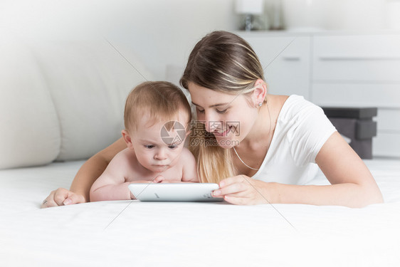 微笑的母亲与婴儿躺在床上并使用数字平板电脑的肖像微笑年轻母亲与婴儿躺在床上并使用数字平板电脑的肖像图片