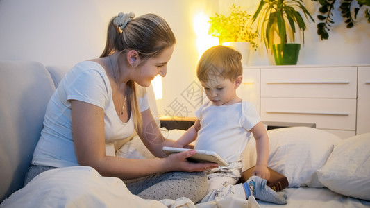 快乐的母亲在睡觉前数字平板上向幼儿子展示卡通快乐的母亲在睡觉前向幼儿子展示数字平板上的卡通图片
