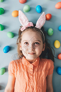 长着粉红兔子耳朵的可爱女孩躺在地板上周围是多彩蛋复活节图片