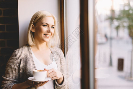 站在窗边的年轻女人拿着一杯咖啡看外面笑自然的户外灯光站在窗边的女士拿着一杯咖啡图片