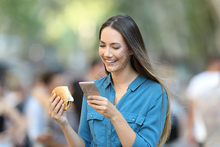 女人拿着汉堡检查智能手机在街上图片