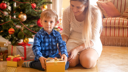 奇迹男孩快乐的幼儿男孩母亲坐在圣诞树下看着礼物和快乐的幼儿男孩微笑的母亲坐在圣诞树下看着礼物和背景