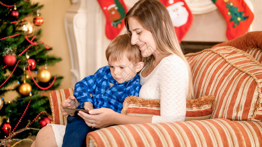 快乐的妈与婴儿在圣诞树旁边的手椅子上观看智能手机视频图片