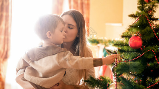美丽的女士亲吻着她孩子而他正在装饰圣诞树美丽的年轻女士亲吻着她的孩子图片