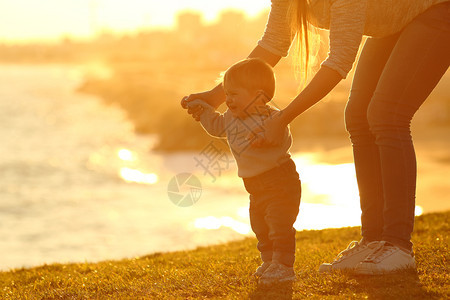 一个孩子的侧面观学习走路和母亲在日落时帮助他在野外的草地上图片