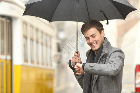 快乐的优雅男子以智能手表咨询信息等待在雨天伞下老镇的街道上等待公共交通图片