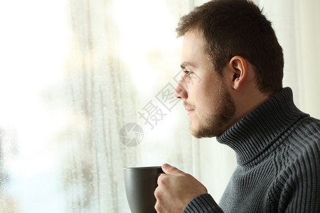 一位满意的男子在雨天透过窗户在家里举着咖啡杯的图片