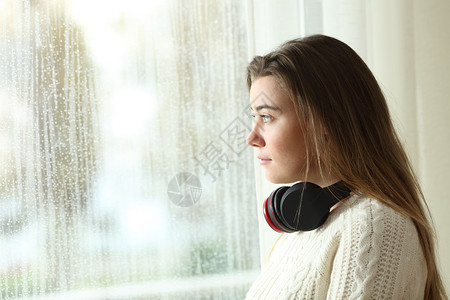 在雨天透过窗户观看一个带着耳机的悲伤青少年侧面肖像图片