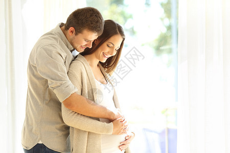 骄傲的新父母看着怀孕亲的肚子站在家里的窗户旁边图片