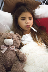 一个小生病的女孩躺在一张床上盖着毯子贴在她最喜欢的玩具熊旁边嘴里有电子温度计图片