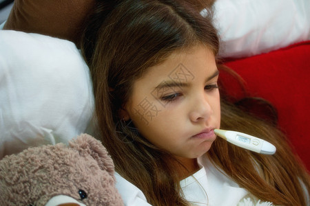一个小生病的女孩躺在一张床上盖着毯子贴在她最喜欢的玩具熊旁边嘴里有电子温度计图片