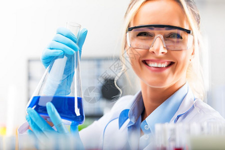 在化学研究实验室内带着保护眼镜和手套拿着含蓝色液体物质的瓶子护目镜和手套年轻有吸引力的年轻女科学家快乐年轻有吸引力的女科学家图片