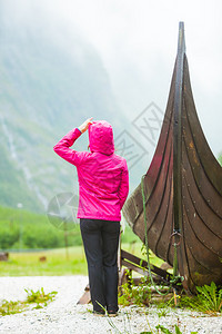 在挪威自然界的旧木型维京船附近站立的女旅游者看着迷雾山雨日旅游和概念在挪威自然界的旧木型维京船附近站立的旅游者图片