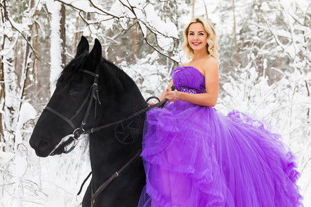 身穿长的粉红裙子在雪冬林骑着黑马的年轻金发美女图片