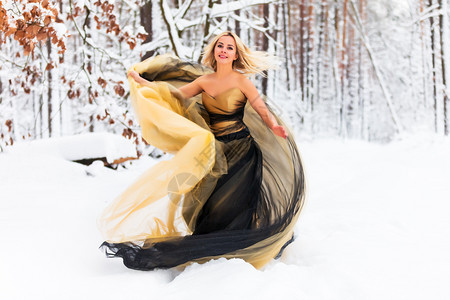 穿着长的黑黄色金裙子的发美女在雪的冬季森林或户外的木中图片