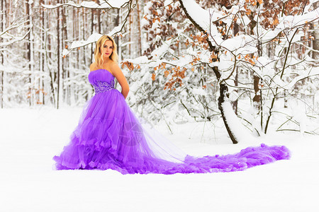 穿着长紫色裙子的金发年轻美女在雪的冬季森林或户外的木里图片
