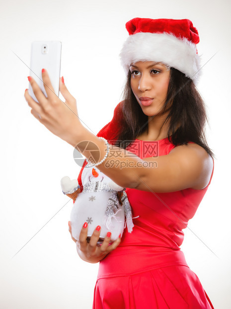 冬季和圣诞节时间概念穿着圣诞帮手帽的妇女穿着红礼服的混血种族女孩带着快乐的雪人玩具用智能手机相拍自图片
