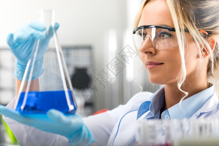 在化学研究实验室内带着保护眼镜和手套拿着含蓝色液体物质的瓶子护目镜和手套年轻有吸引力的年轻女科学家快乐年轻有吸引力的女科学家图片