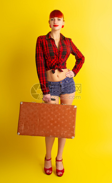 穿着结的格子衬衫和短裤的漂亮女孩带着一个黄光背景的复古手提箱图片