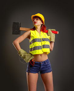 一个穿黄色头盔手套的女孩穿着短裤黄头盔毛手套的建筑背心在黑暗景上拿着一把大锤子图片