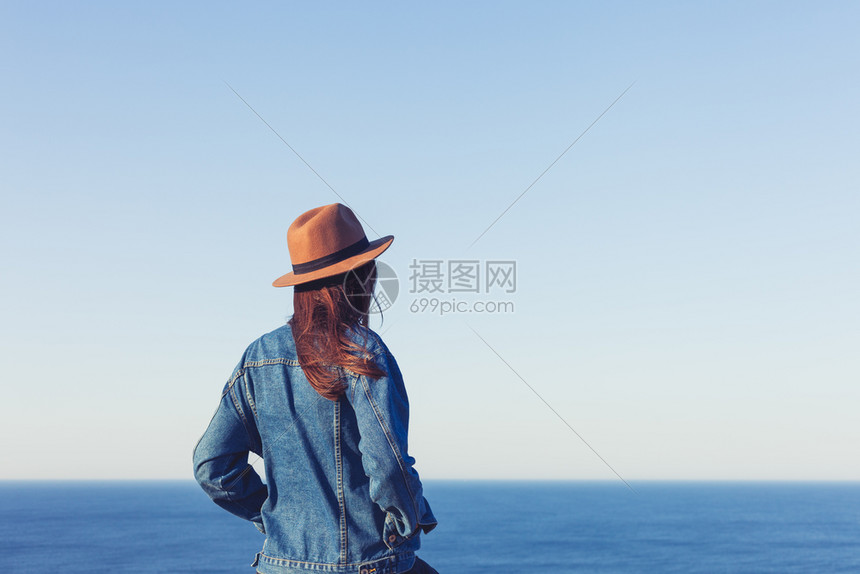 穿着牛仔裤夹克和帽子站着看蓝色的海洋和天空图片