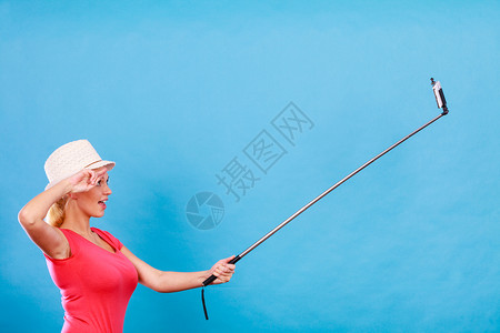戴太阳帽的女旅游客用她的智能手机在自拍棒上照工作室摄的是蓝背景旅行女金发游客用电话拍照图片