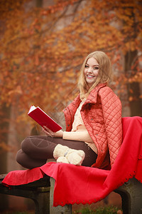 秋天在公园里穿着夹克的微笑妇女坐在长椅上手持书籍穿着夹克微笑的妇女在公园里坐着图片