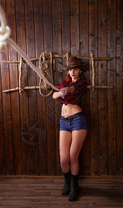 身着传统牛仔服装的年轻女孩穿着宽厚的帽子和一件格衬衫在黑暗的房间里扔着套索图片