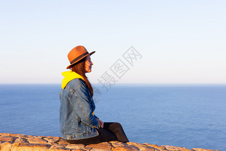 穿旅行服装和戴帽子的妇女坐着看蓝海和天空旅行概念照片坐着和看蓝海天空的妇女图片