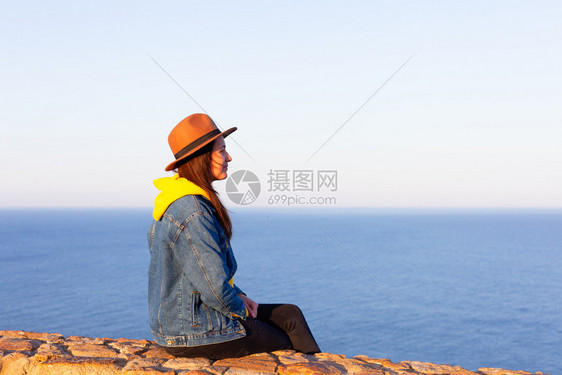 穿旅行服装和戴帽子的妇女坐着看蓝海和天空旅行概念照片坐着和看蓝海天空的妇女图片