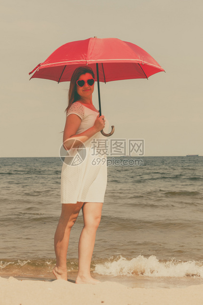 红发妇女穿着带纱布的白裙子在海滩上举着红色雨伞在沙滩上走来去在暑假散步时享受舒适的放松红发妇女在海滩上走来去在海滩上举着雨伞图片