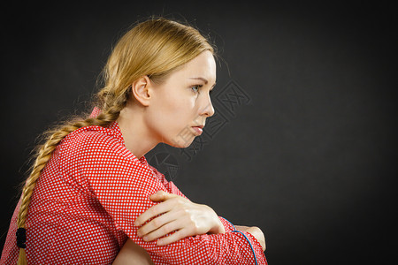 患有抑郁症的年轻女子金发孩穿着红色睡衣的令人沮丧年轻女子,穿着红色睡衣膝盖围着双臂深黑的墙背景。图片