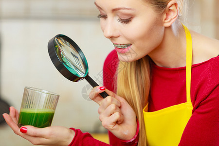 厨房的年轻女子拿着绿色健康的蔬菜冰凉果汁杯透过放大玻璃寻找图片