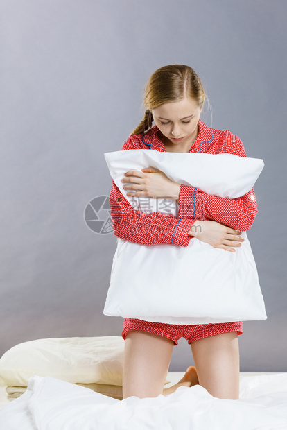 患有抑郁症的年轻女子金发孩穿着红色睡衣坐在枕头的床上穿着灰墙背景的色底图片