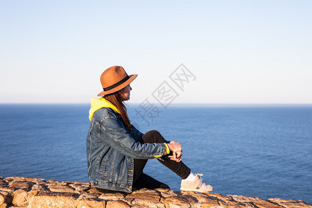 穿旅行服装和戴帽子的妇女坐着看蓝海和天空旅行概念照片坐着和看蓝海天空的妇女图片