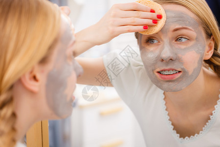 年轻女照镜子洗手间除去面部粘土罩和海绵女用除去泥面罩图片