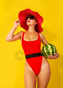 年轻女孩穿着明亮的红色泳衣和宽的红帽子在黄色背景上吃西瓜图片